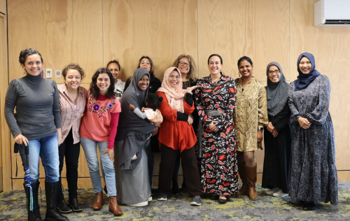 Twelve women of different ages and ethnicities stand in a row posing for a photo. One is holding a young child, The women are smiling, with arms around each other, looking into the camera's lens. 