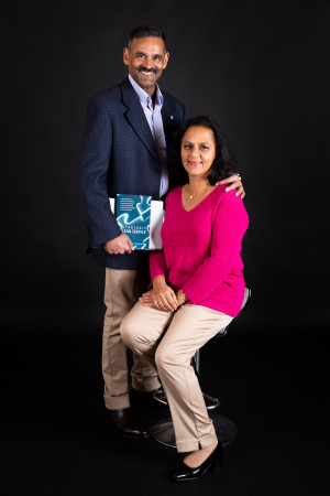 Man and woman posing for a photo. The man is standing up, with his hand on the woman's shoulder. She is sitting down. The man holds a blue book.