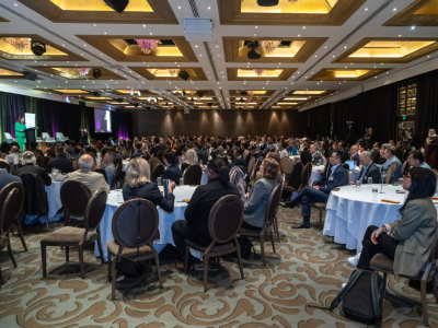 A photo of a conference room with hundreds of people seated around tables, looking to the stage where a person is speaking